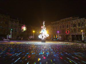 Photo of a town square with a fir tree in disco balls in the center illuminating the ground with multiple colors