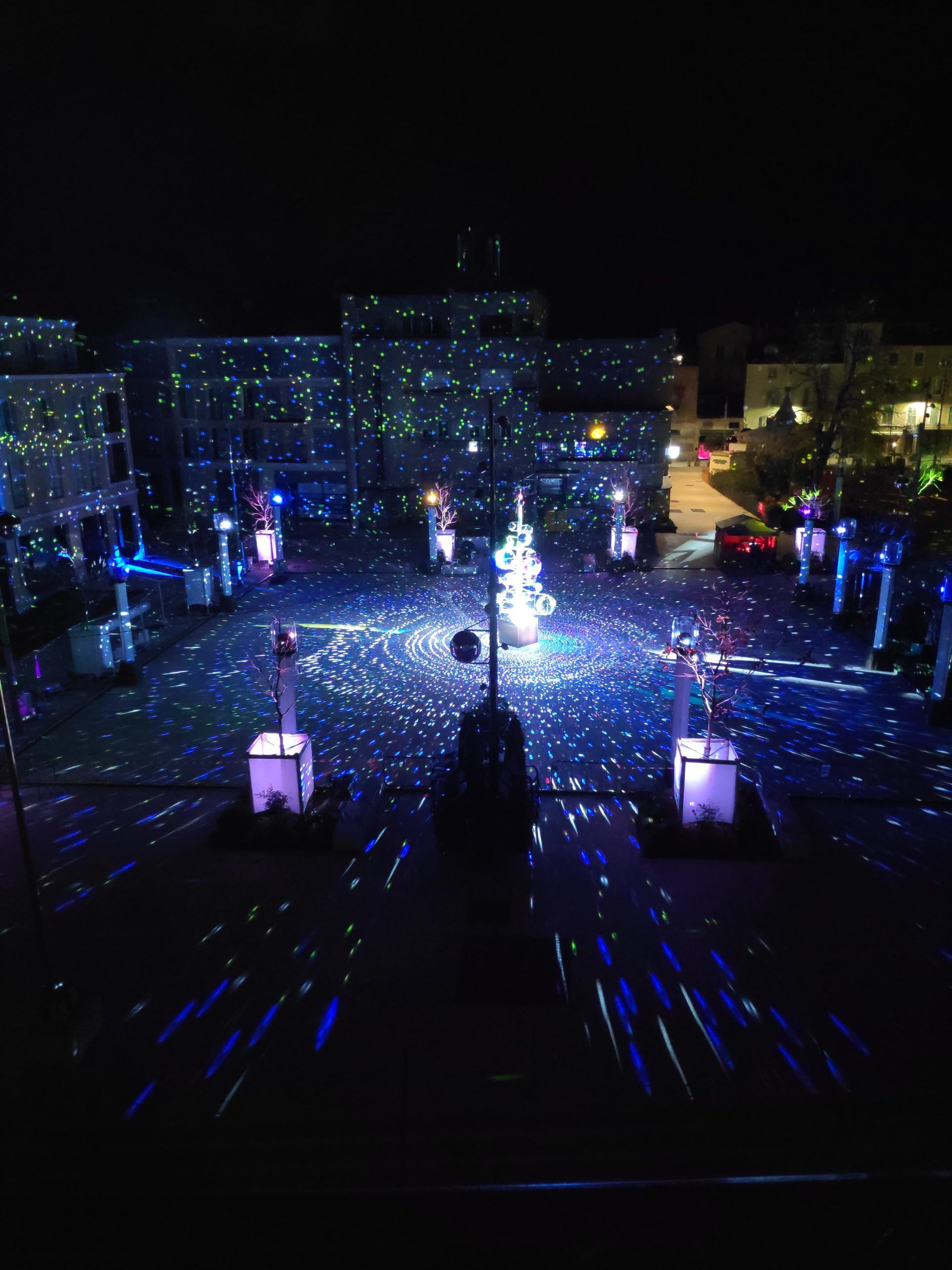 Photo of a town square with a tree and white, blue and green lights in Antibes
