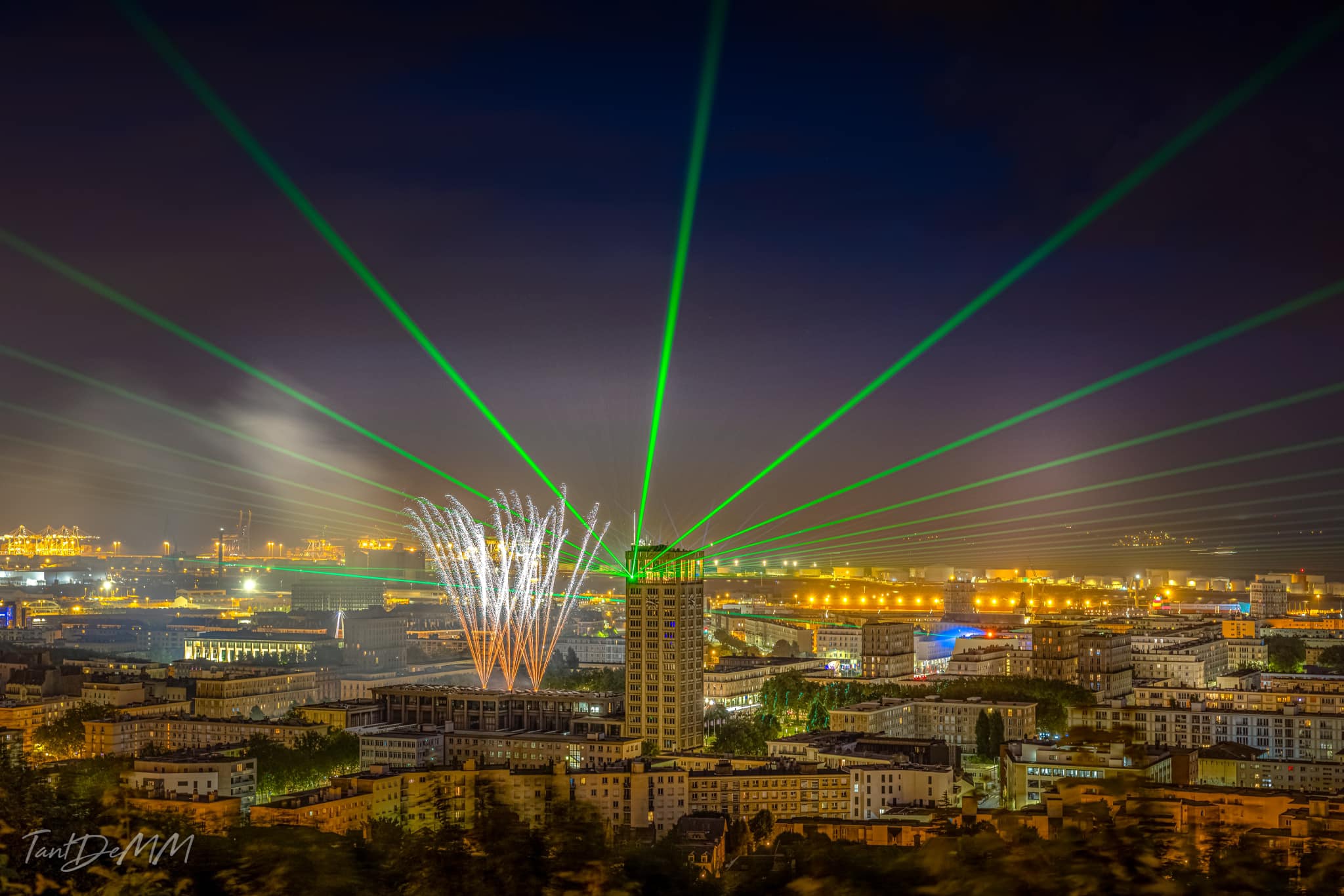 Photo of a town tower with green laser beams projection starting from the tower and accompanied by fireworks