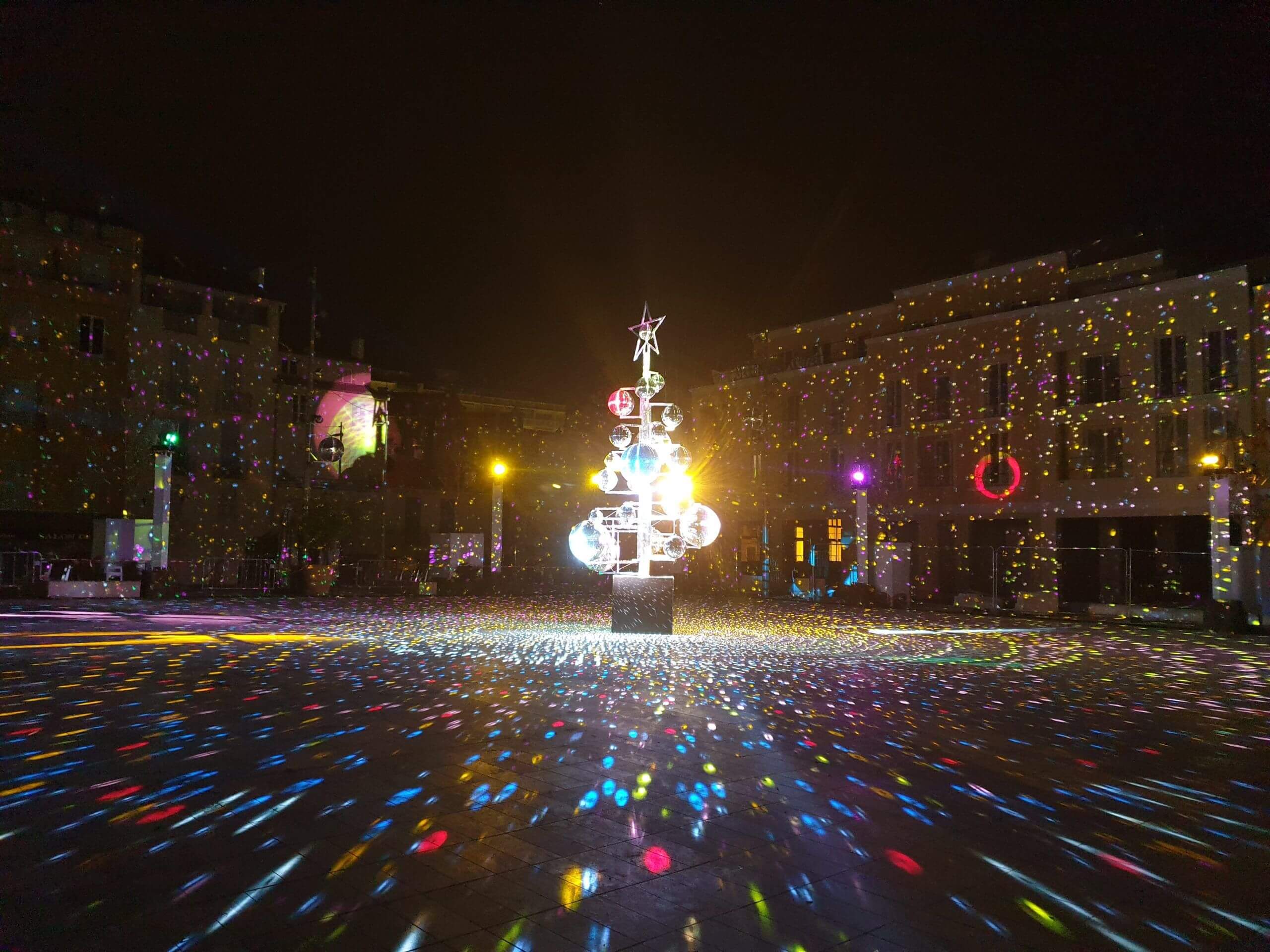 Europe Évènement - Photo d'une place avec un arbre aux lucioles au centre illuminant le sol de couleurs multiples